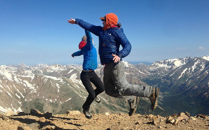Two people jump in the air and high five with a mountainous landscape in the background